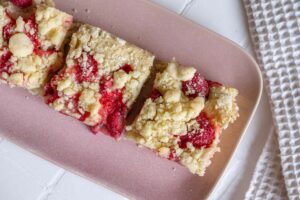 Einfacher Erdbeer-Streuselkuchen vom Blech: Drei quadratische Stücke Streuselkuchen mit Erdbeeren unter der Streuselmasse liegen auf einem rosa Teller. Dieser steht auf einem weißen, gekachelten Untergrund.