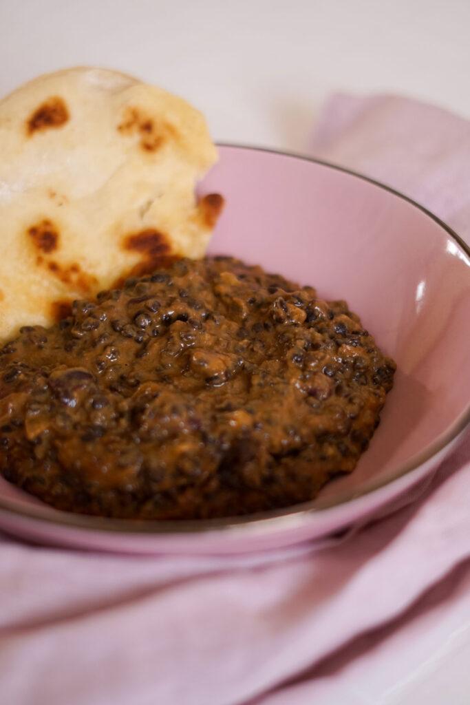 Indisches Linsengericht Dal Makhani auf einem rosa Teller mit Pfannenbrot im Hintergrund