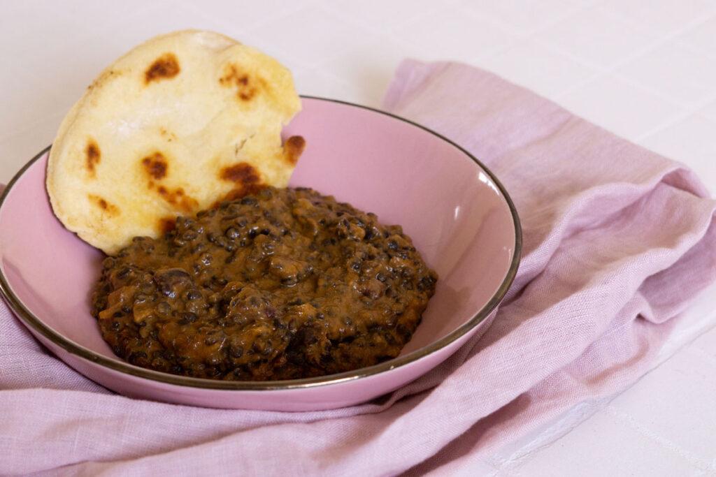 Indisches Linsengericht Dal Makhani auf einem rosa Teller mit Pfannenbrot im Hintergrund