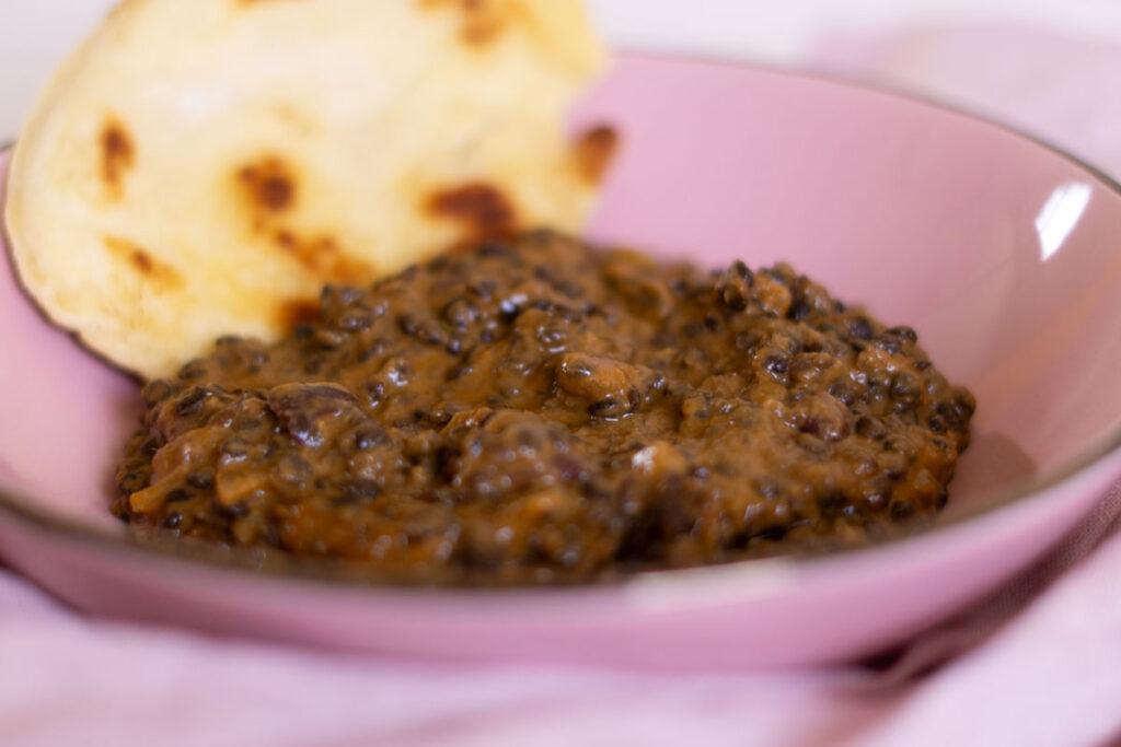 Indisches Linsengericht Dal Makhani auf einem rosa Teller mit Pfannenbrot im Hintergrund